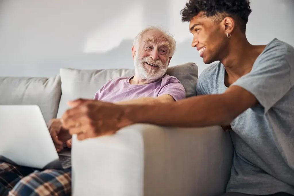 A man sitting on a couch with an older man, both engaged in conversation and smiling at each other.