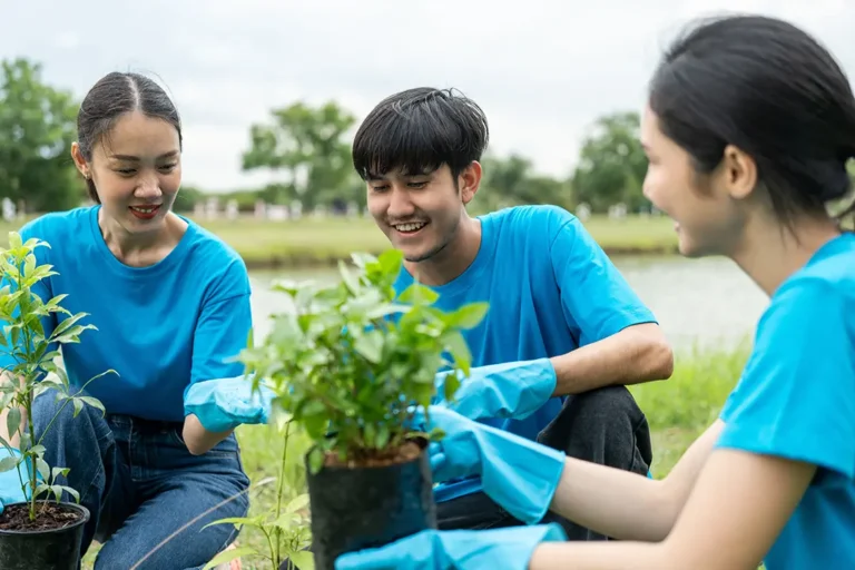 Three young individuals in blue shirts and gloves are planting trees in a green outdoor setting.